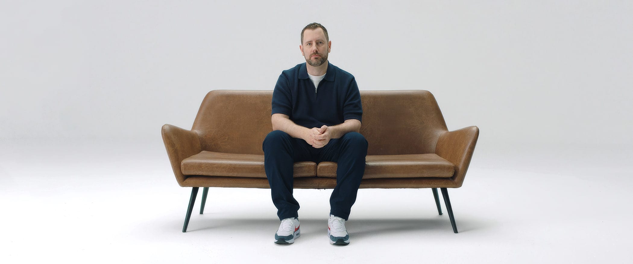 Joshua Davies sitting on a leather sofa against a plain pale background, hands together, looking at the camera.