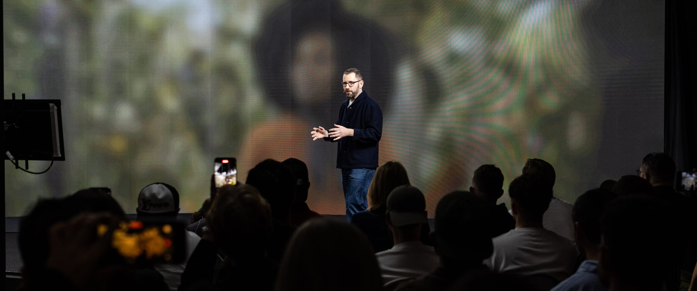 Joshua Davies on stage mid-gesture, a large screen behind him, the backs of a seated audience in the foreground.