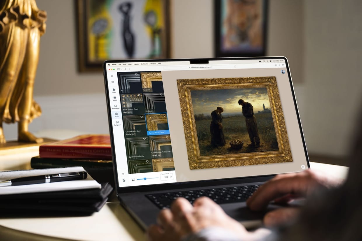 A laptop on a desk in a gallery office running FrameEdit: a scrolling library of frame profiles down one side, and a nineteenth-century painting of two figures in a field shown inside a carved gilt frame. Framed works hang on the wall behind.