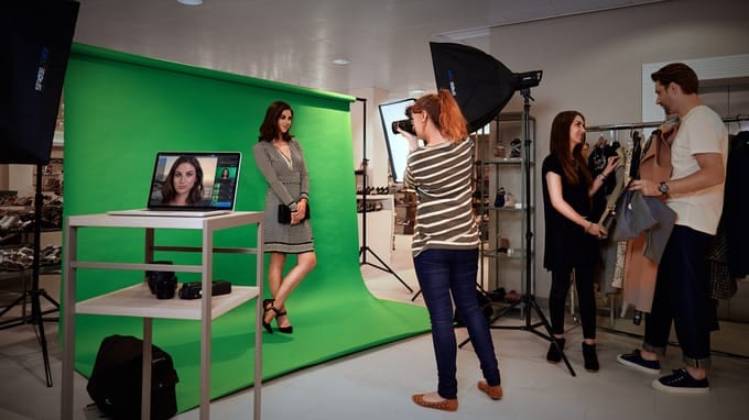 A PhotoKey shoot in a clothing shop: a model against a green backdrop, a photographer working handheld, and a laptop on a stand beside them already showing the composited result.