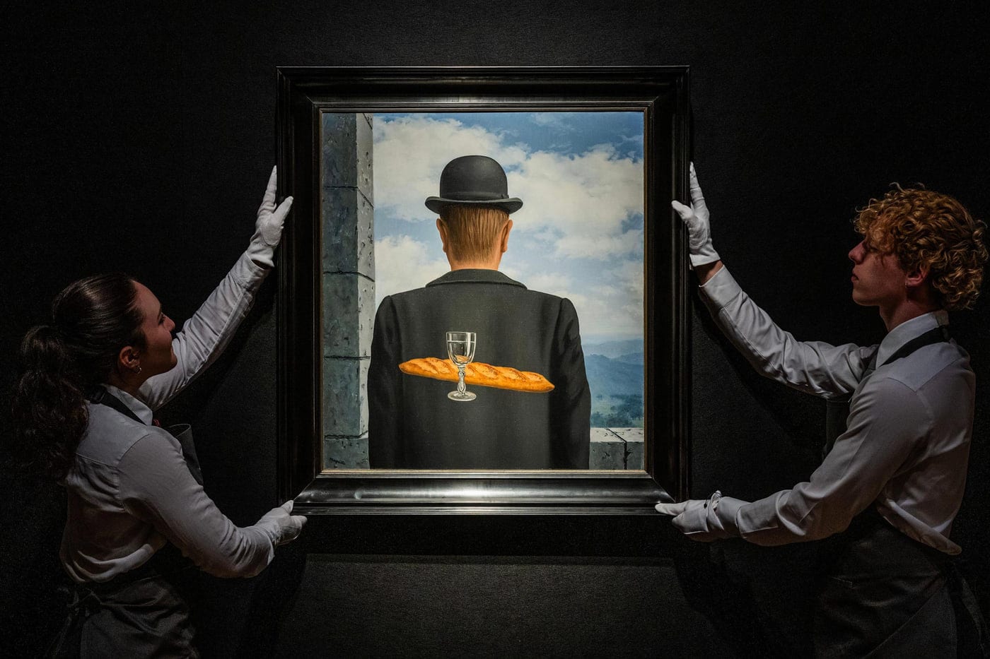Two gallery technicians in white gloves holding a framed Magritte against a black wall: a man in a bowler hat seen from behind, with a baguette and a glass of water in front of him, against a cloudy sky.
