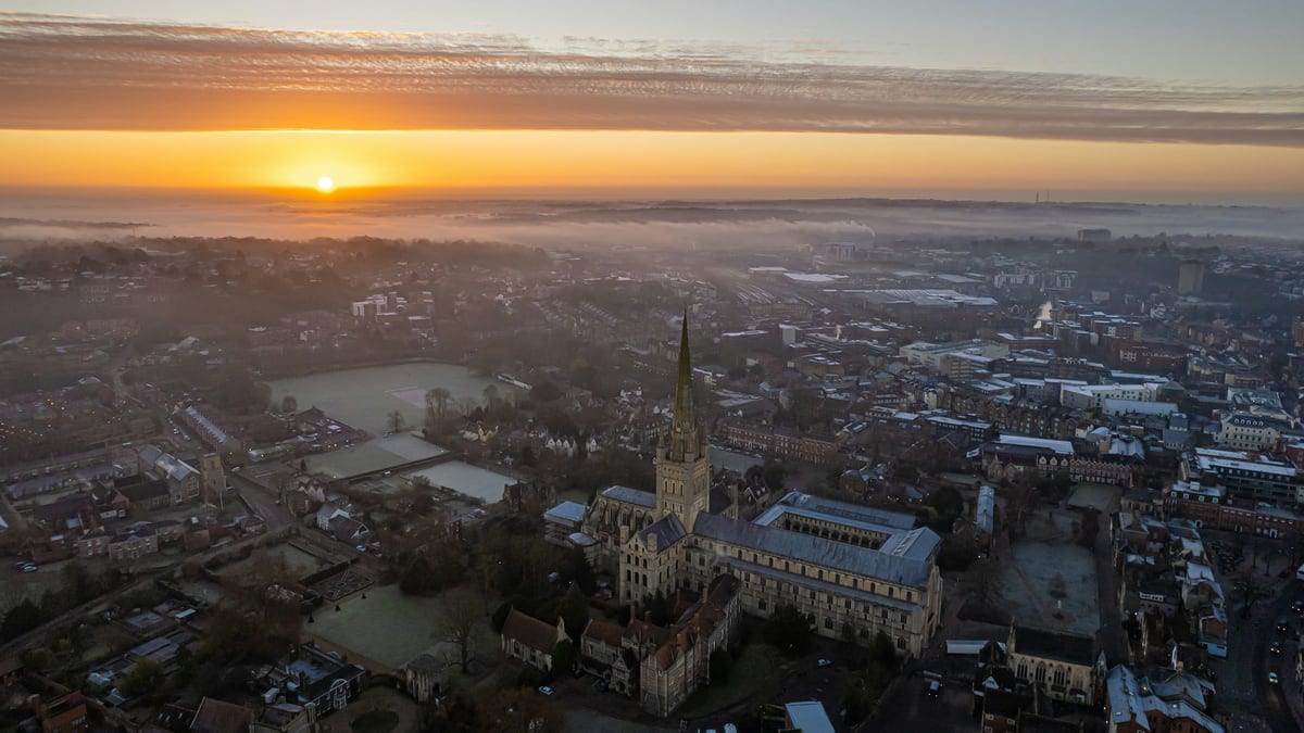 An aerial view of Norwich: the cathedral and its spire standing above the rooftops, frost on the greens around it, mist lying across the city, and a low sun beneath a long band of cloud.