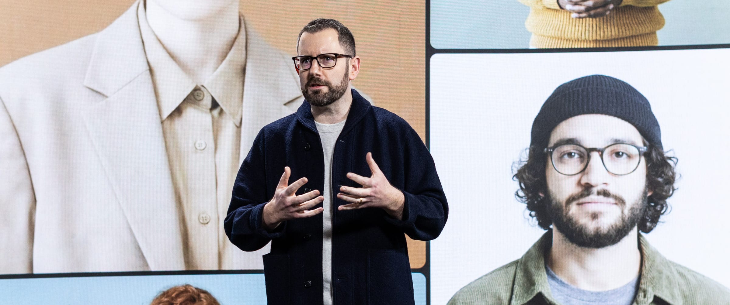 Joshua Davies speaking, mid-gesture with both hands, in front of a wall-sized screen of portraits of different people.